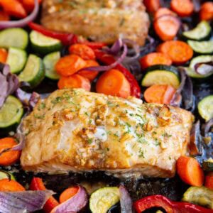 Cooked cod filet next to roasted vegetables on a baking sheet with another cooked cod filet in the background.