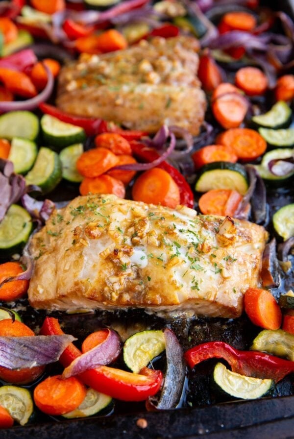 Cooked cod filet next to roasted vegetables on a baking sheet with another cooked cod filet in the background.
