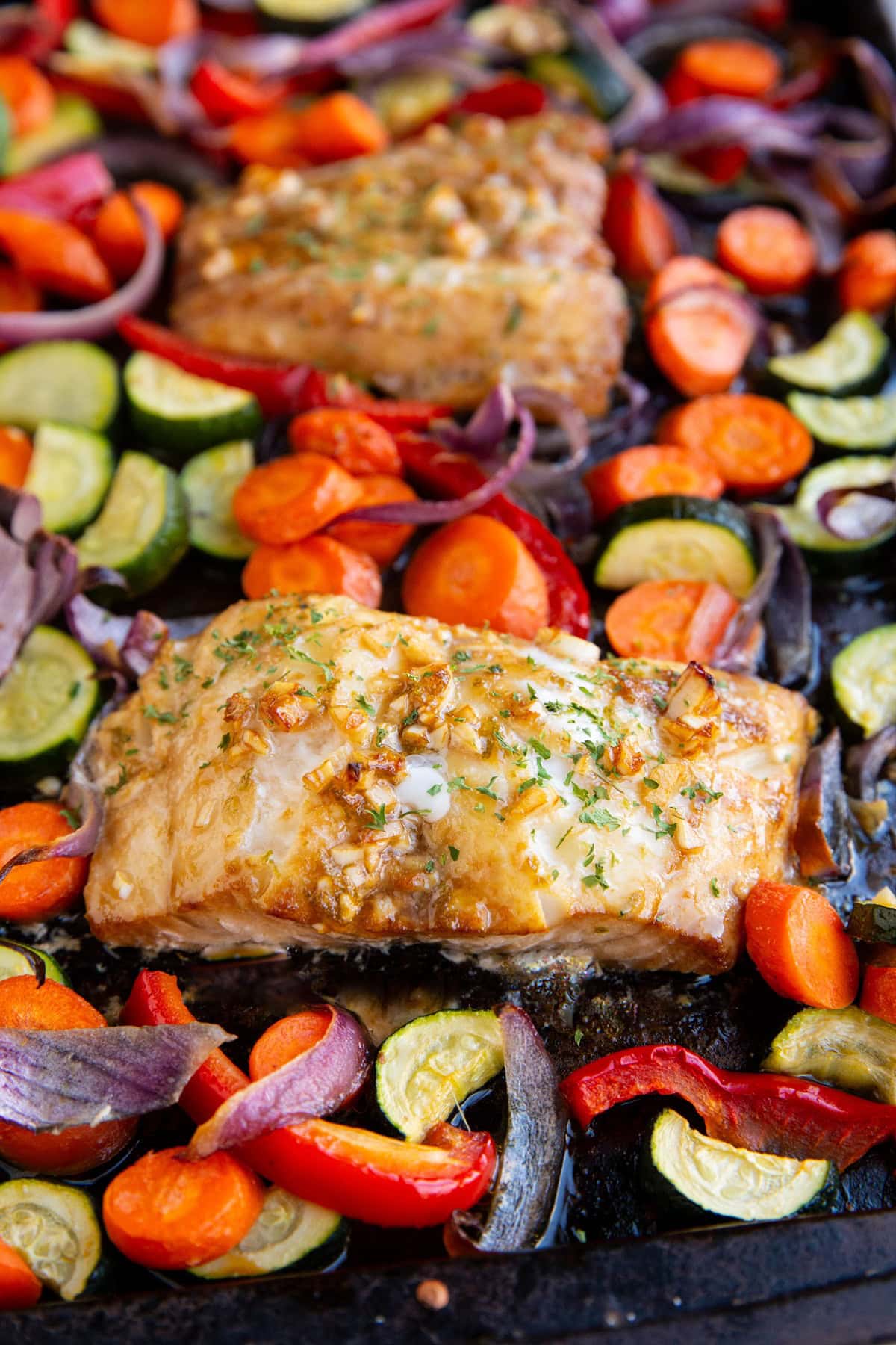 Cooked cod filet next to roasted vegetables on a baking sheet with another cooked cod filet in the background.