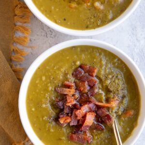 Two white bowls full of split pea soup with crispy cooked bacon on top and a golden napkin to the side, ready to serve.