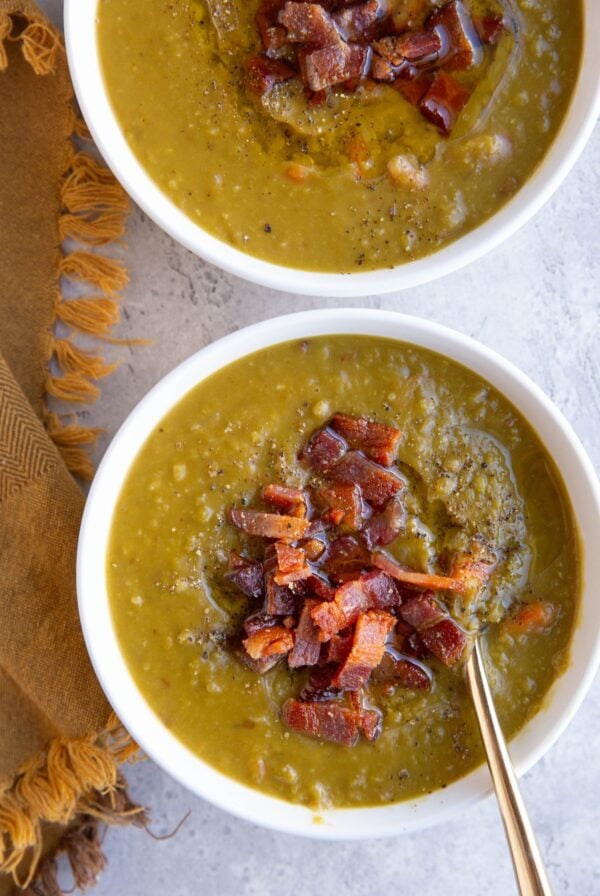 Two white bowls full of split pea soup with crispy cooked bacon on top and a golden napkin to the side, ready to serve.