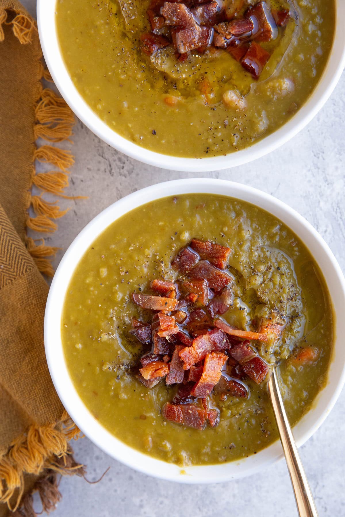 Two white bowls full of split pea soup with crispy cooked bacon on top and a golden napkin to the side, ready to serve.