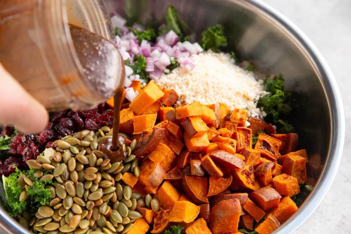 Stainless steel bowl full of salad ingredients with a hand pouring balsamic vinaigrette over the salad to dress it.