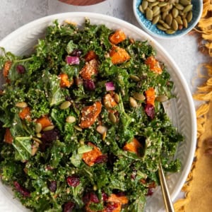 large white serving bowl filled with kale and sweet potato salad with a small bowl of pumpkin seeds, a small bowl of dried cranberries, and a golden napkin to the side.