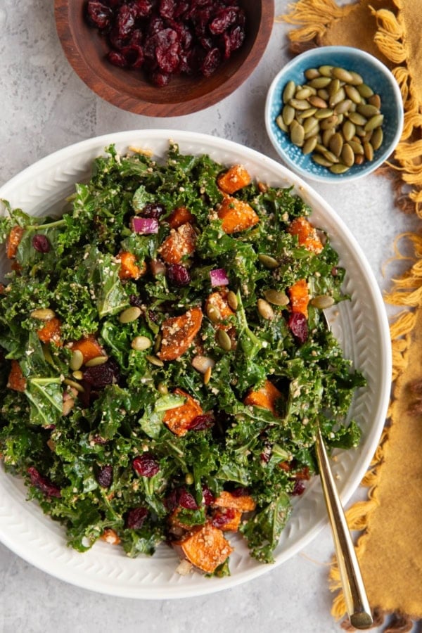 large white serving bowl filled with kale and sweet potato salad with a small bowl of pumpkin seeds, a small bowl of dried cranberries, and a golden napkin to the side.