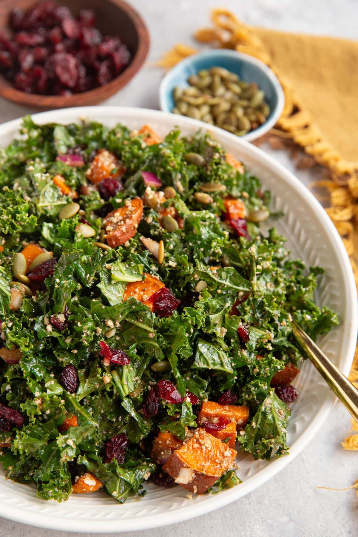 large salad bowl full of kale and sweet potato salad with a golden serving spoon to the side. A wooden bowl of dried cranberries and a blue bowl of pumpkin seeds are in the background with a golden napkin.