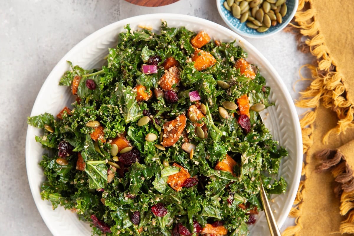 Sweet potato kale salad in a large white bowl with a golden serving spoon and a golden napkin to the side.