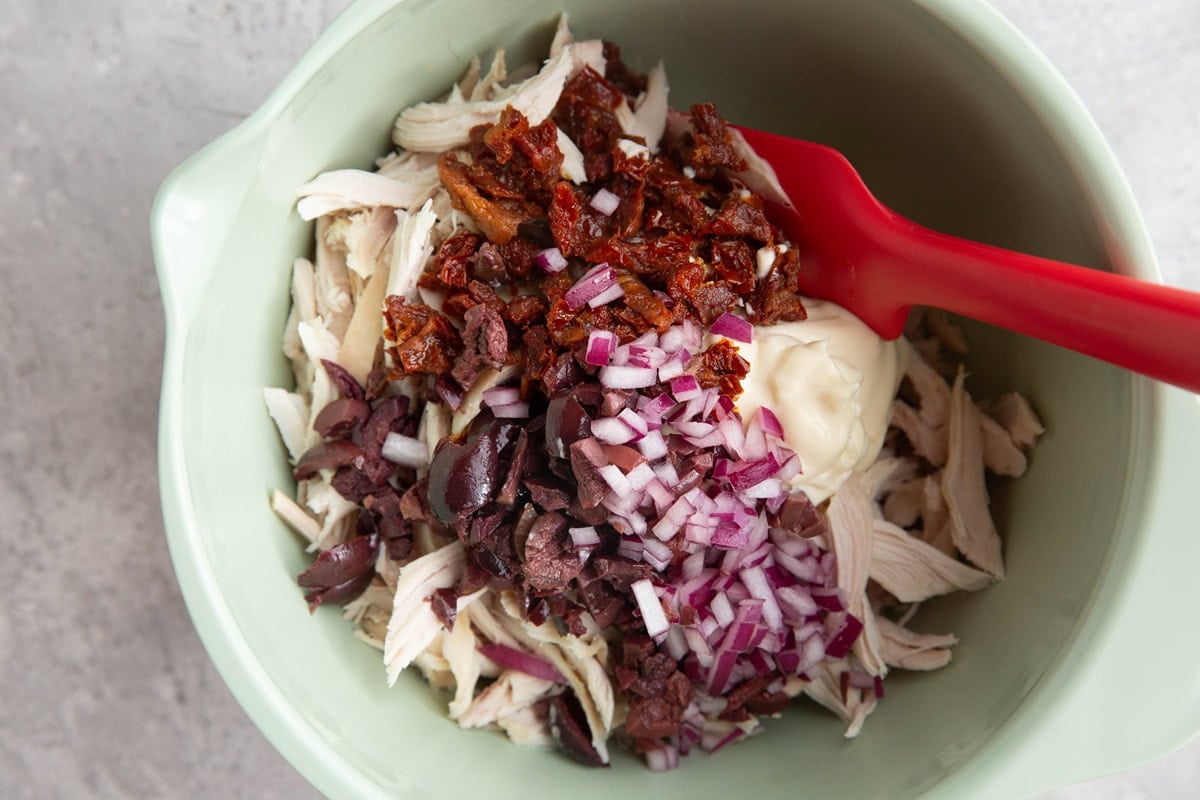 Mixing bowl of rotisserie chicken, mayonnaise, chopped red onions, sun-dried tomatoes, and kalamata olives with a rubber spatula