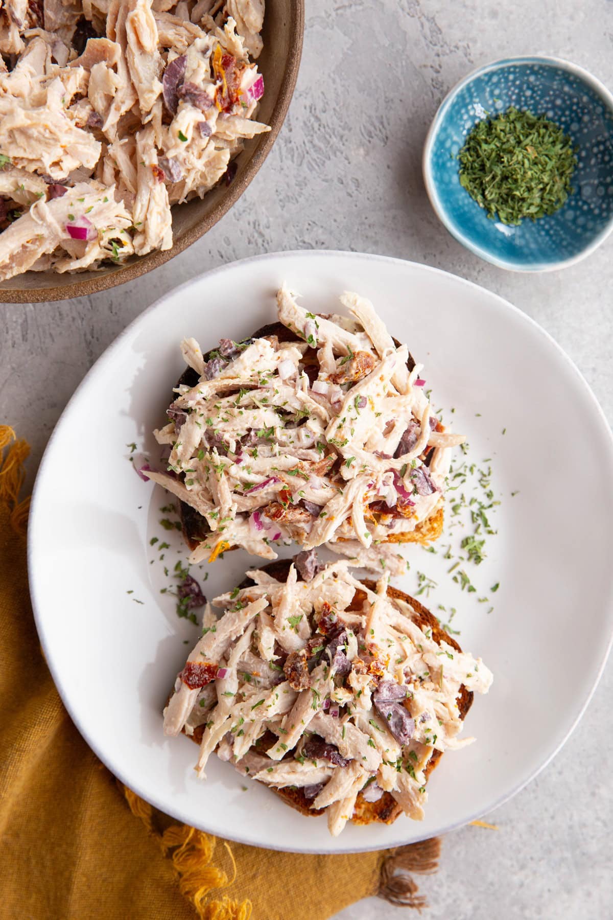 Two slices of toast topped with chicken salad on a white plate and the bowl with the rest of the chicken salad to the side.