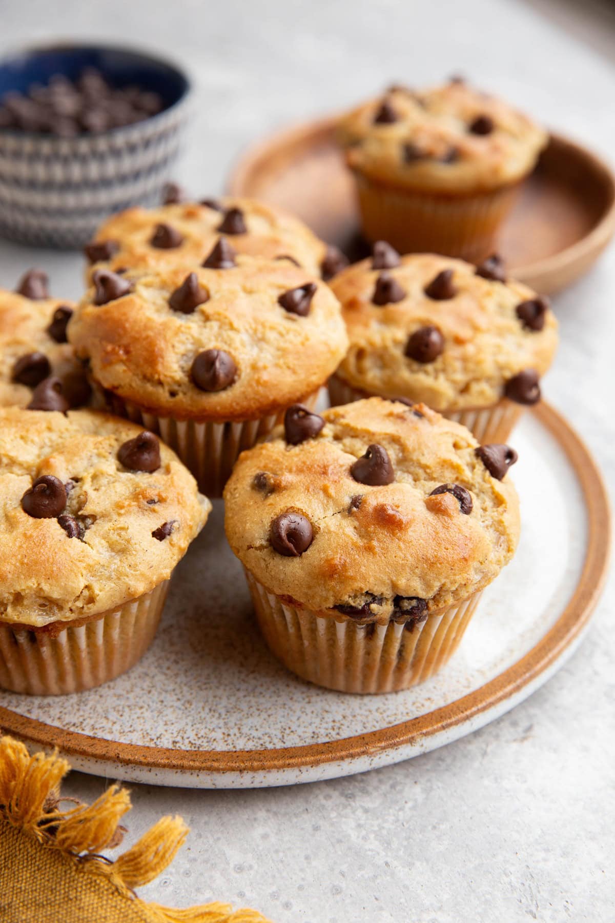Plate of chocolate chip muffins with a smaller wooden plate with a single muffin in the background and a bowl of chocolate chips.