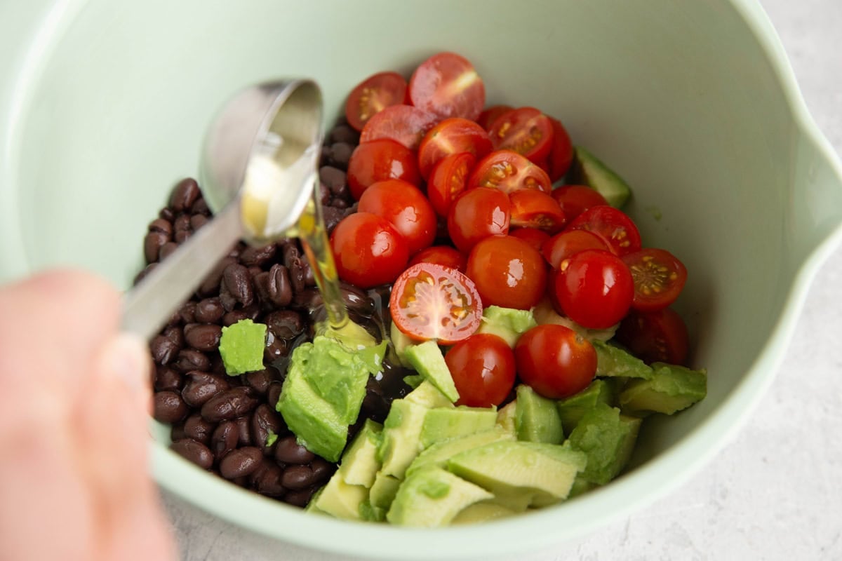 Mixing bowl full of black beans, avocado, and cherry tomatoes with a hand pouting oil into the bowl for dressing.