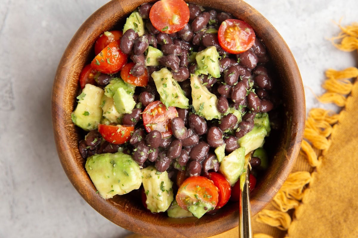 Wooden bowl full of black bean and avocado salad with a gold serving spoon and a golden napkin to the side.