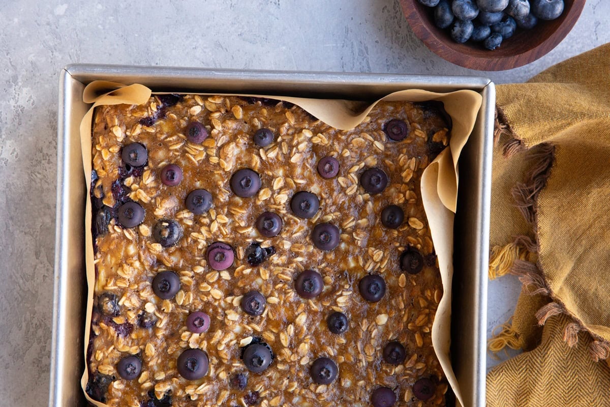 Baking dish with blueberry breakfast bake with a golden napkin and a bowl of fresh blueberries.