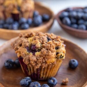 Two wooden plates with blueberry buttermilk muffins and fresh blueberries. A bowl of fresh blueberries in the background.