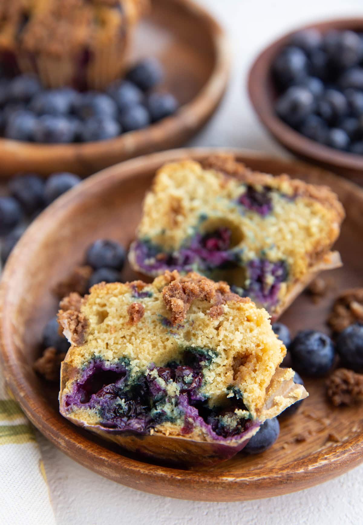 Blueberry muffin sliced in half on a wooden plate with another plate of blueberry muffin in the background and a small bowl full of fresh blueberries.