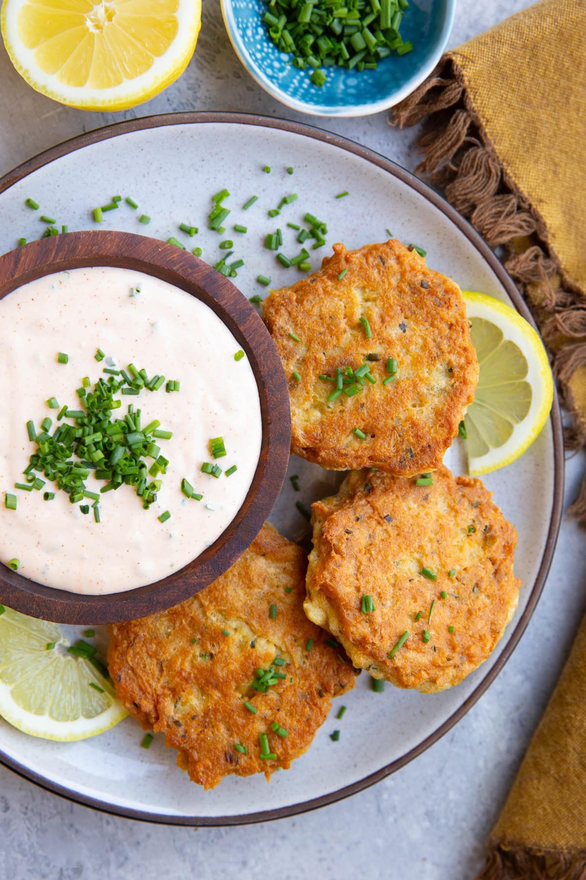 Three golden brown salmon patties on a plate with a bowl of like pink sauce to the side, resembling crab cake sauce with lemon slices on the plate and a bowl of chives to the side.