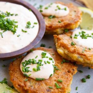Salmon patties on a plate drizzled with crab cake sauce and sprinkled with chopped chives with the bowl of sauce to the side and fresh lemon slices.