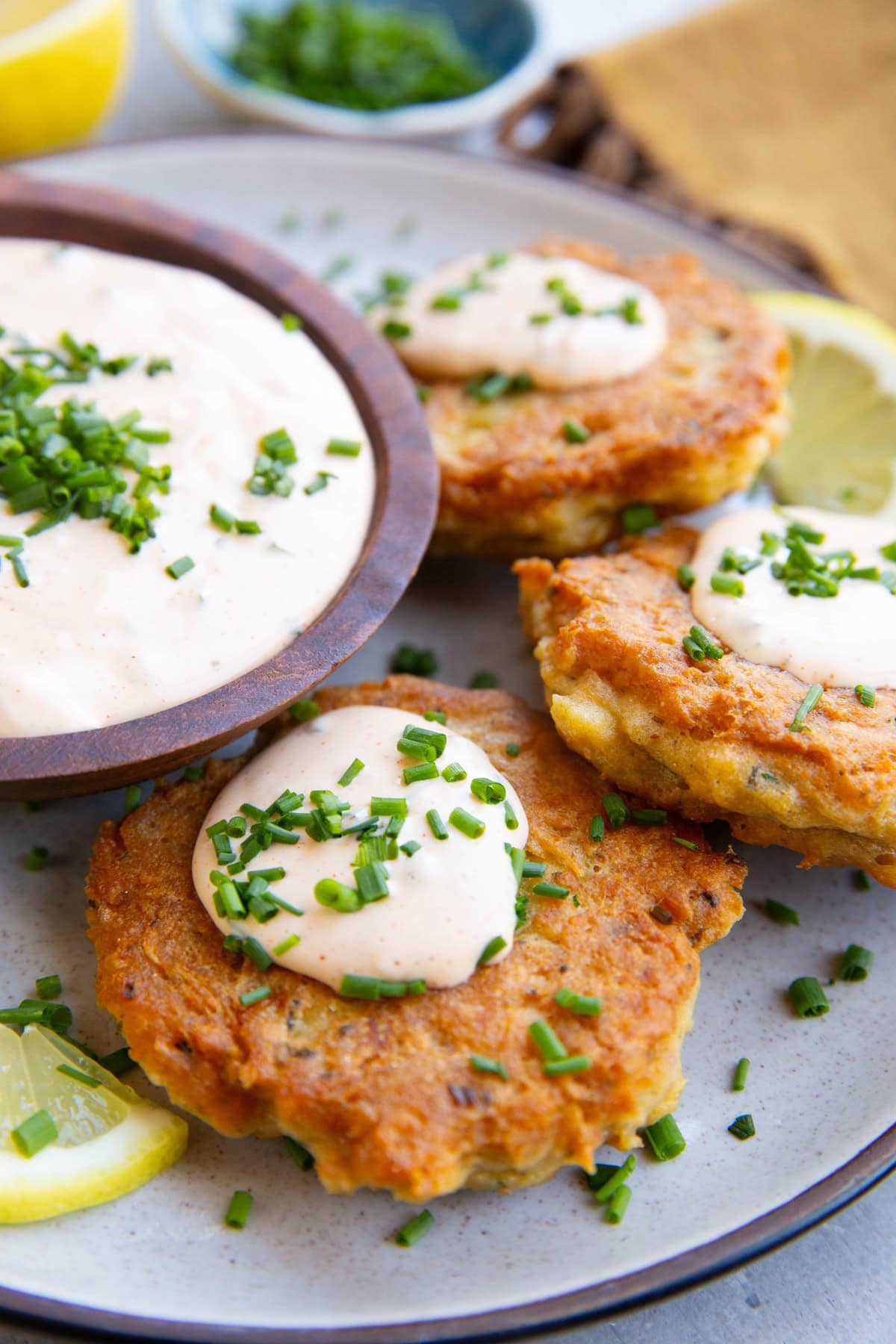 Salmon patties on a plate drizzled with crab cake sauce and sprinkled with chopped chives with the bowl of sauce to the side and fresh lemon slices.