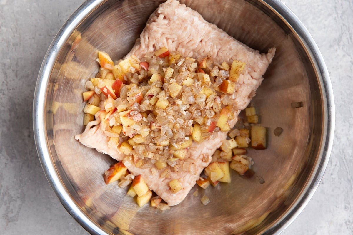 Stainless steel mixing bowl with a pound of raw ground chicken and sauteed onion and apple on top, ready to be mixed together.