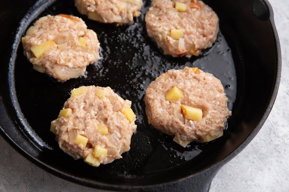 Cast iron skillet with raw chicken sausage patties cooking in oil.