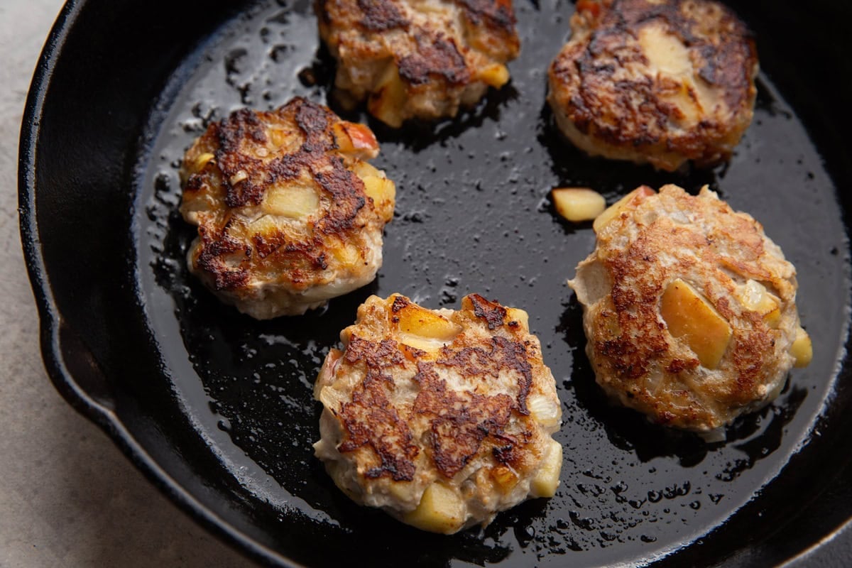 Cast iron skillet with five chicken sausage patties cooking in oil. The tops are golden brown with visible chunks of apples.