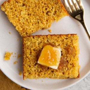 Large slice of cornbread cut in half on a plate with butter and honey on one side and a gold fork, ready to serve.