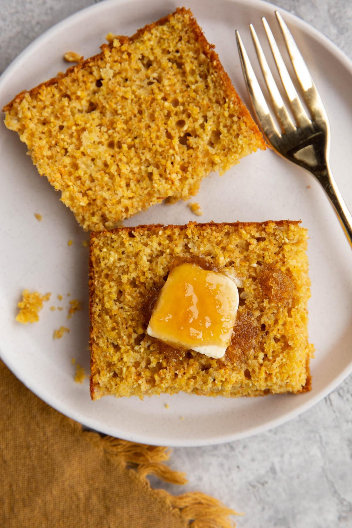 Large slice of cornbread cut in half on a plate with butter and honey on one side and a gold fork, ready to serve.