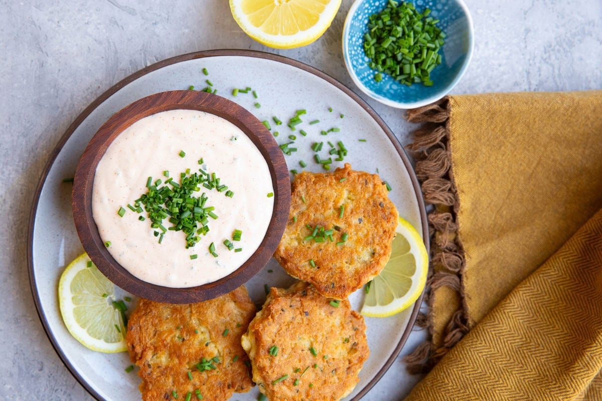 Three salmon cakes on a plate with a wooden bowl full of crab cake sauce and a small bowl of chives to the side with half of a lemon.