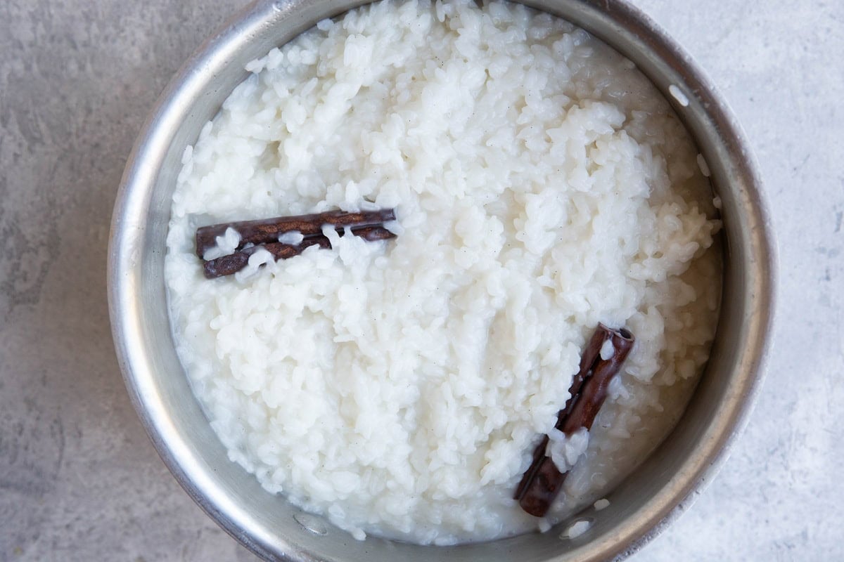 A metal bowl filled with white rice pudding, garnished with two sticks of cinnamon on top. The bowl is placed on a light gray surface.