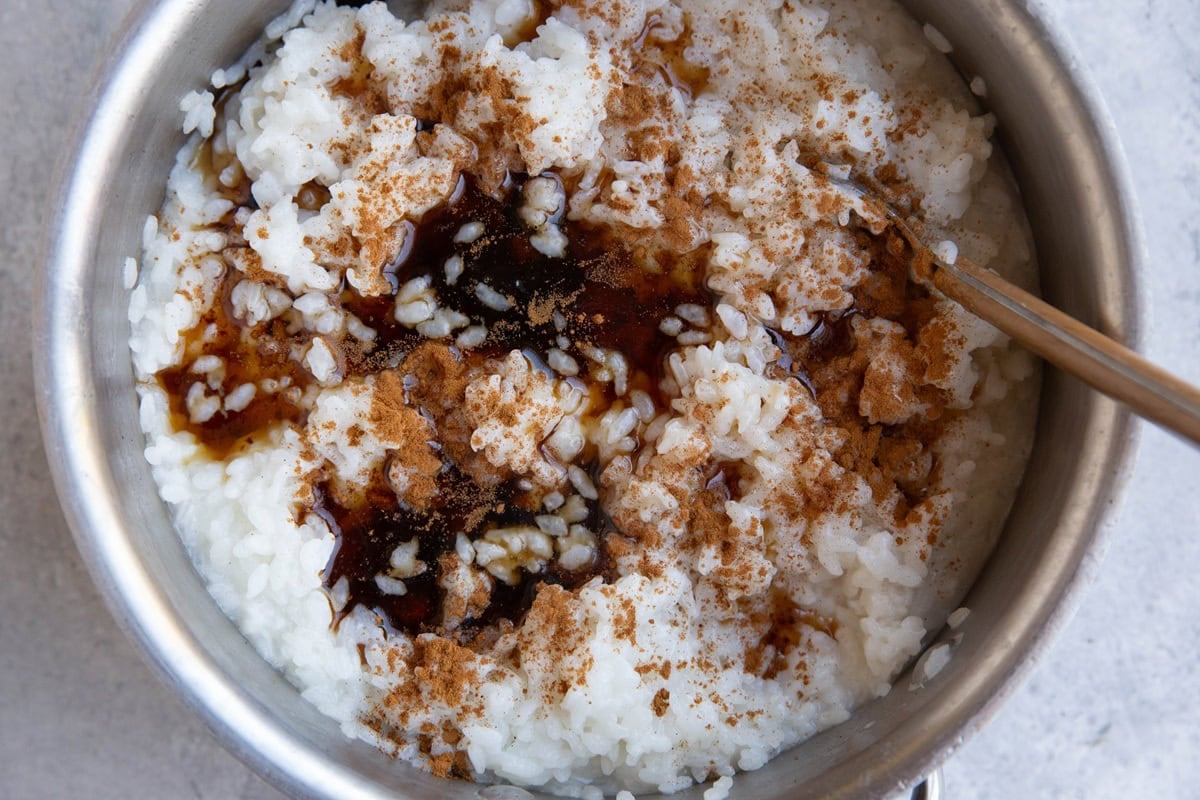 A pot of rice pudding topped with a drizzle of dark syrup and sprinkled with ground cinnamon, with a spoon resting on the side.