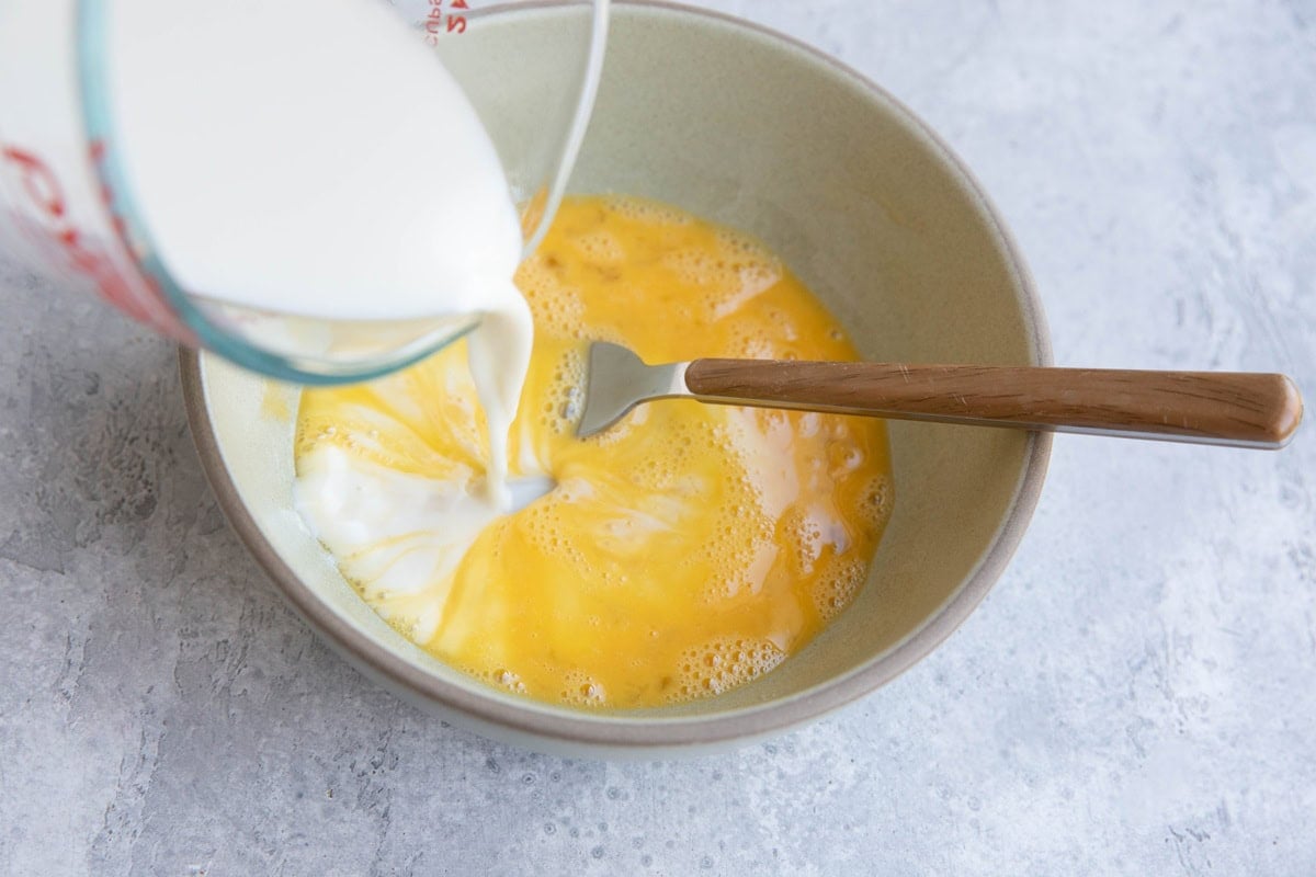 A bowl with beaten eggs is being mixed with milk poured from a measuring cup, and a fork with a brown handle is in the bowl, ready for mixing.