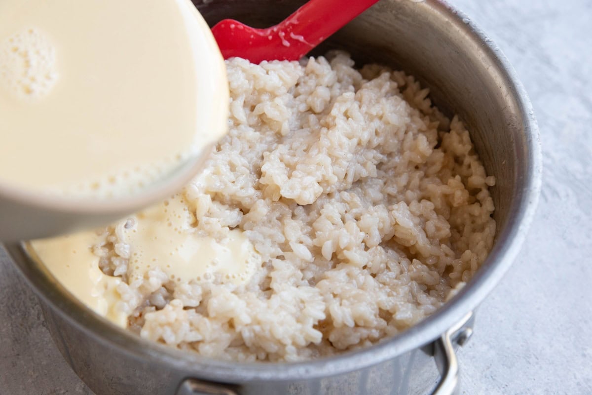 A pot of cooked rice with a creamy liquid being poured over it, next to a red spatula on a light-colored surface.