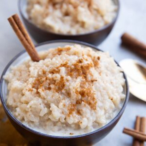 Two bowls of creamy rice pudding topped with ground cinnamon and garnished with cinnamon sticks, sitting on a yellow cloth with a spoon nearby.