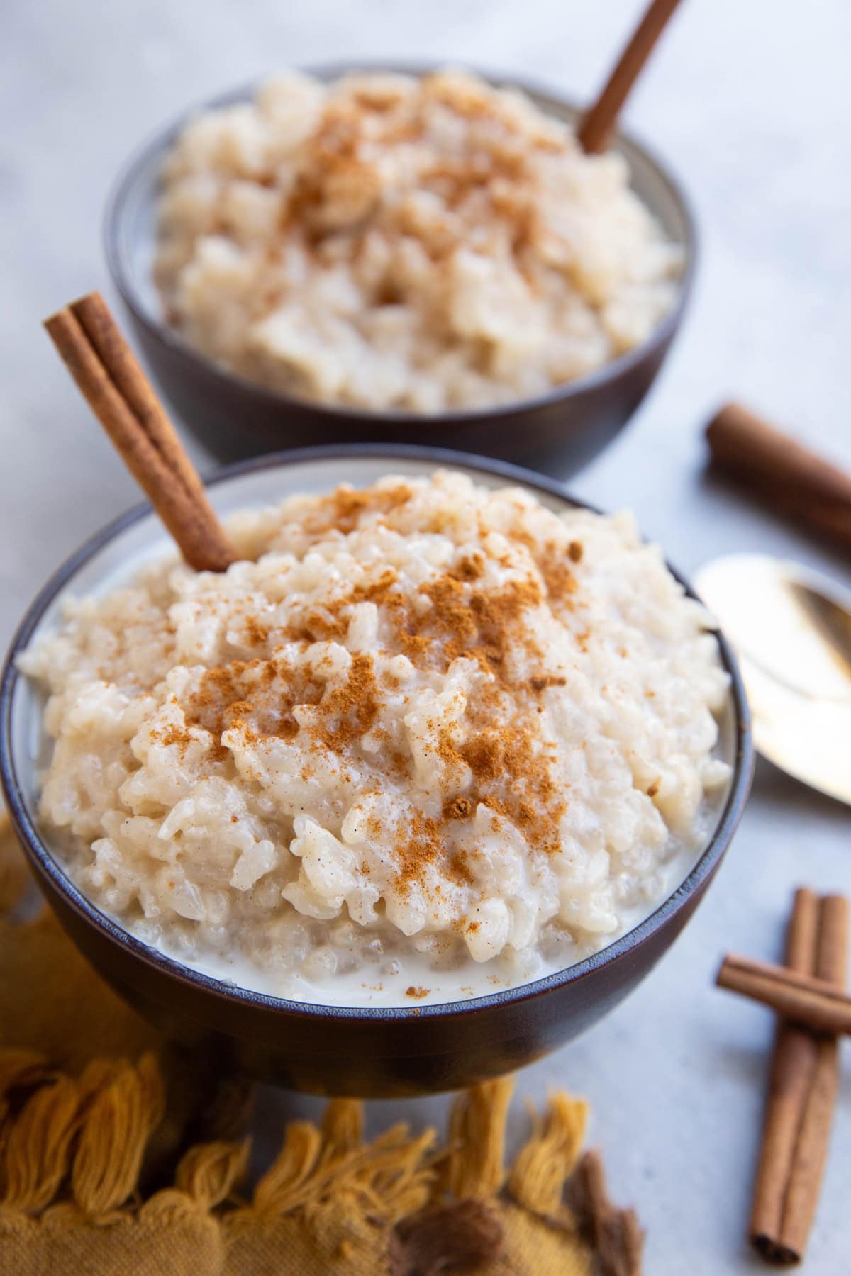 Two bowls of creamy rice pudding topped with ground cinnamon and garnished with cinnamon sticks, sitting on a yellow cloth with a spoon nearby.