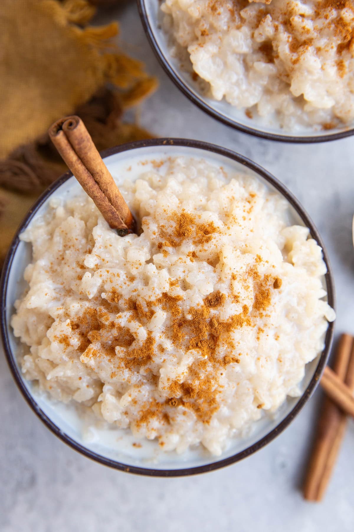 A bowl of creamy rice pudding sprinkled with cinnamon, garnished with a cinnamon stick, on a light surface with a brown cloth nearby.