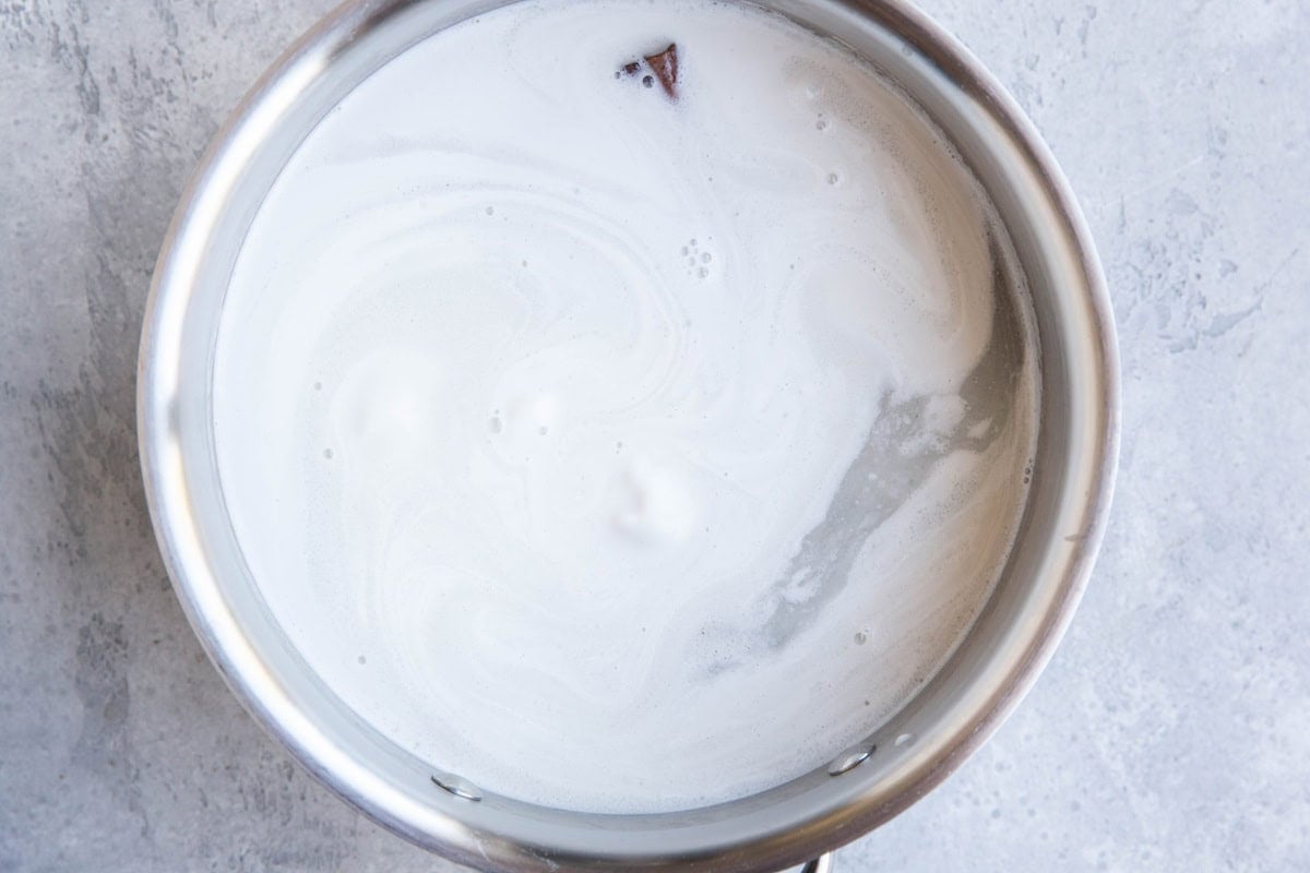 A stainless steel pot filled with simmering milk, with a few bubbles and swirls visible on the surface, sits on a light gray countertop.
