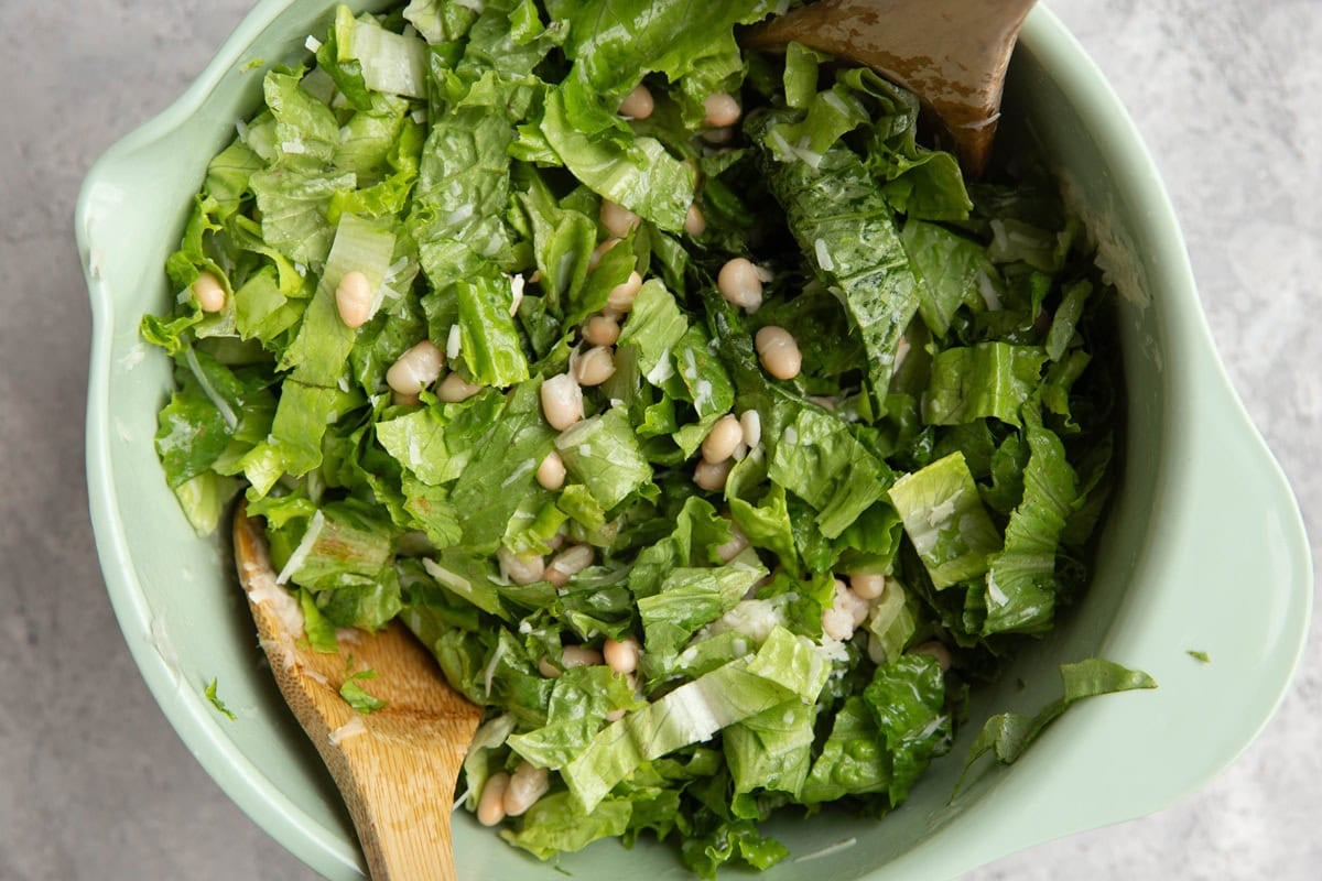 Salad mixed together in a mixing bowl with romaine lettuce, white beans, and parmesan cheese and two wooden spoons to mix the salad.