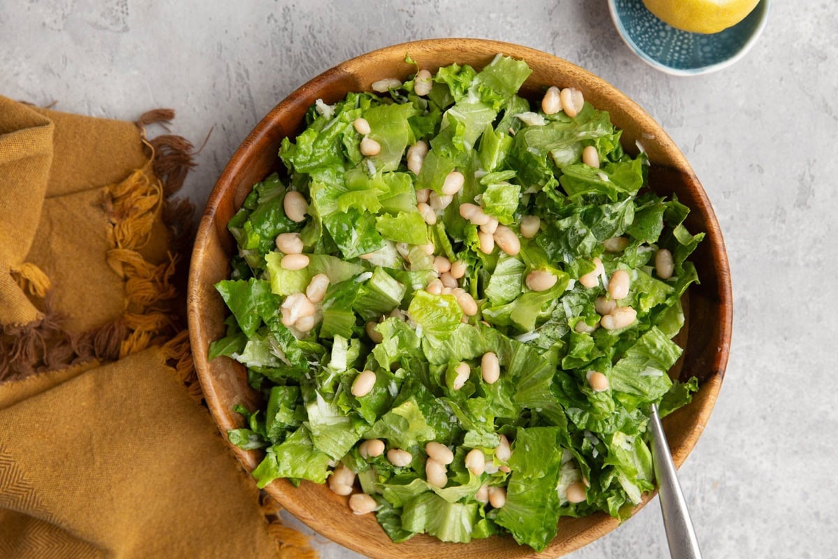 Wooden bowl of white bean lemon parmesan salad with a golden napkin to the side and half of a lemon.