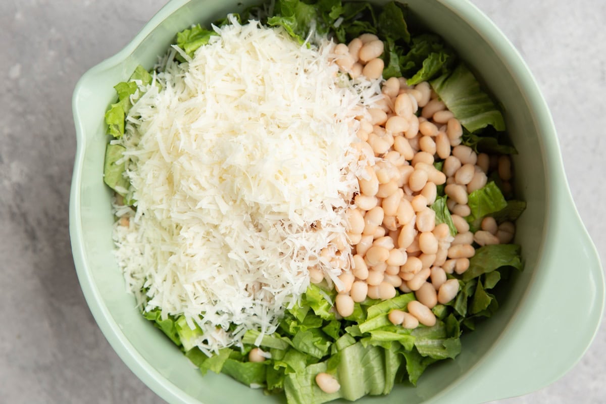 Chopped romaine lettuce, white beans, and grated parmesan cheese in a large mixing bowl, to be made into salad.