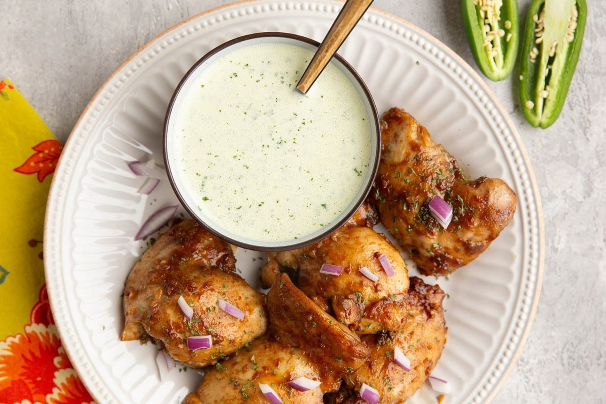 Peruvian chicken on a white plate with a bowl of green sauce and a sliced in half jalapeno to the side.