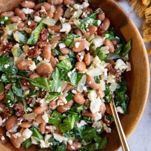 Large wooden bowl full of pinto bean salad with feta cheese, collard greens, and sun-dried tomatoes. A golden spoon in the bowl ready to serve and a golden napkin to the side.