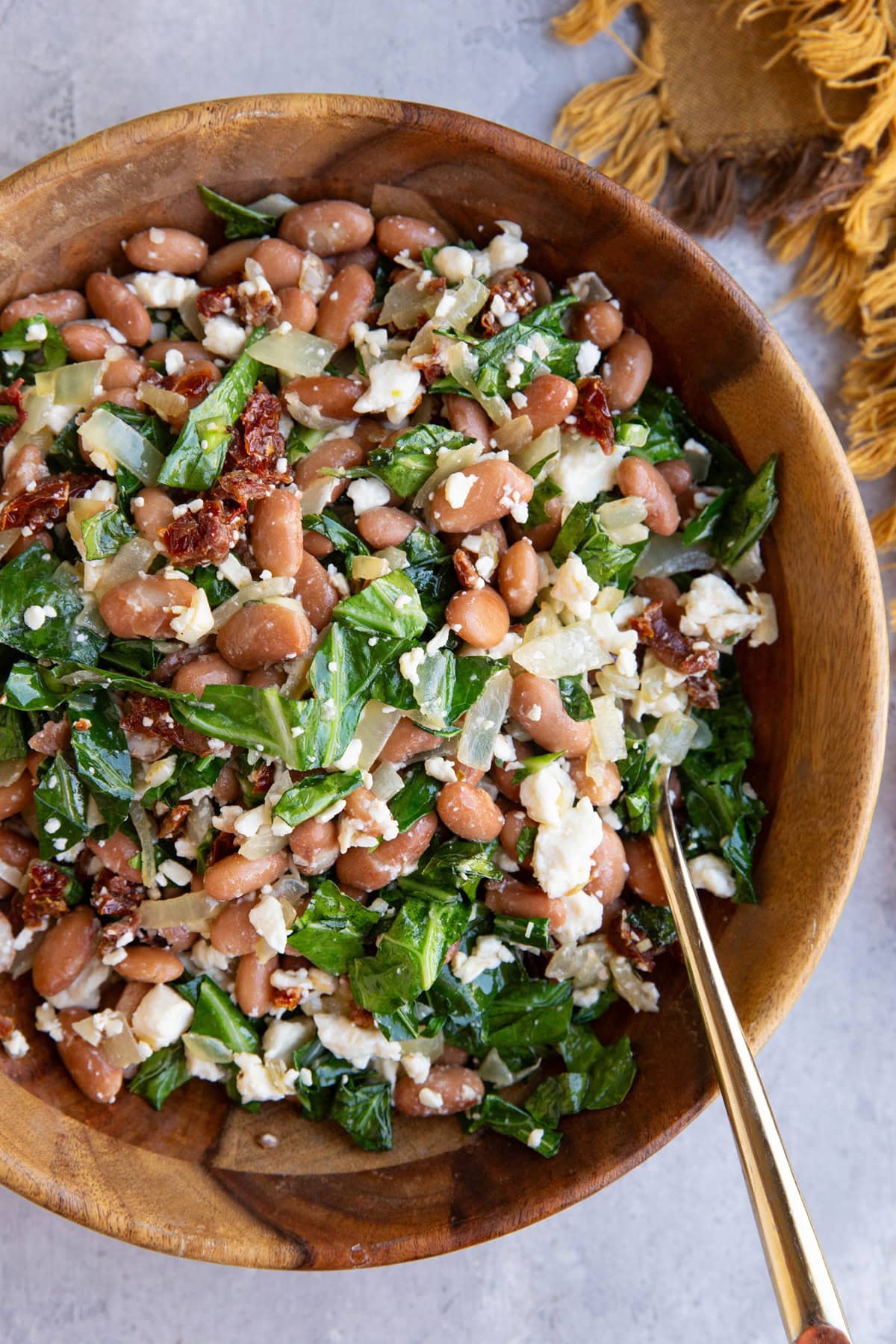 Large wooden bowl full of pinto bean salad with feta cheese, collard greens, and sun-dried tomatoes. A golden spoon in the bowl ready to serve and a golden napkin to the side.