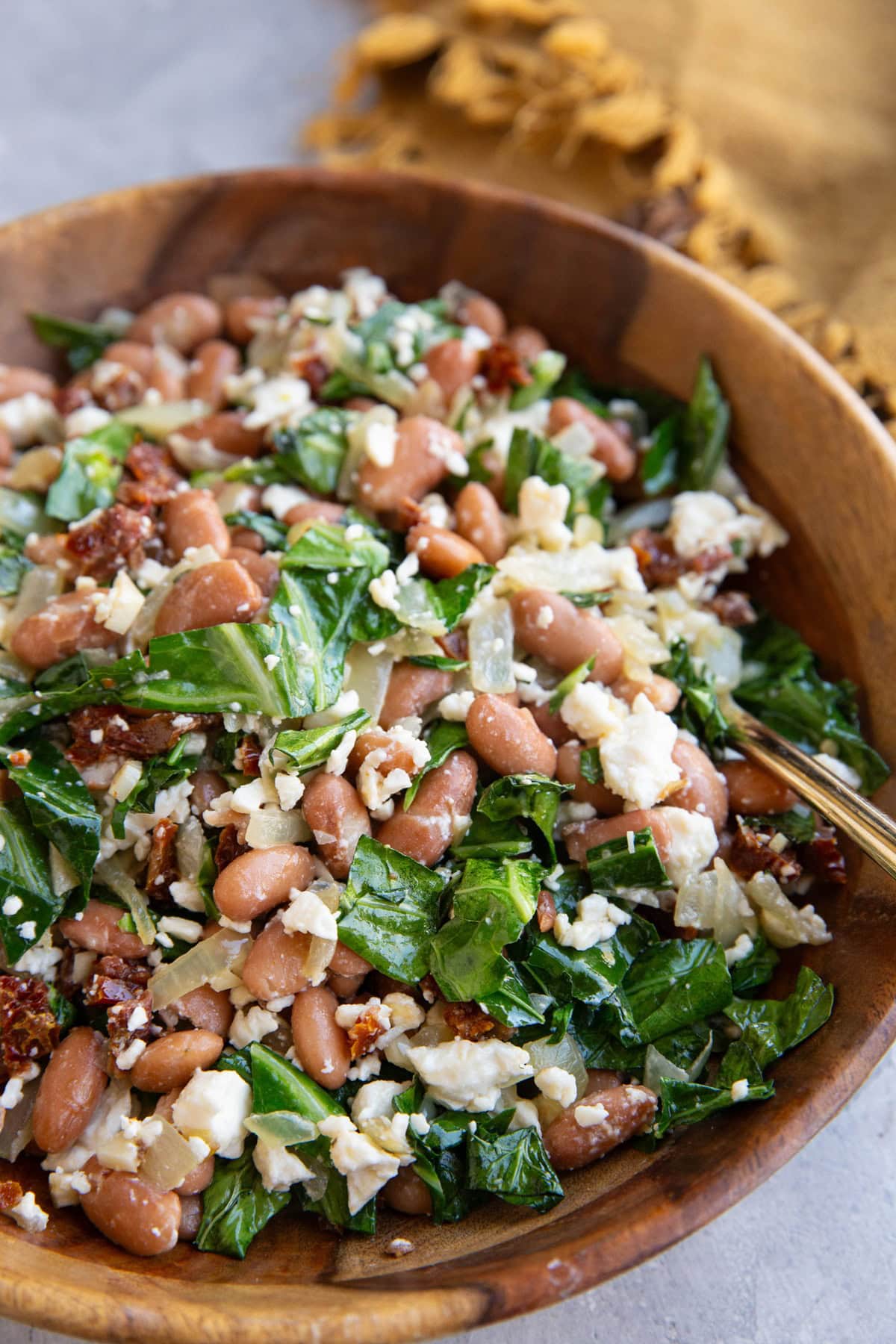 Wooden bowl full of pinto beans, collard greens, feta cheese, and sun-dried tomatoes. An easy side salad.