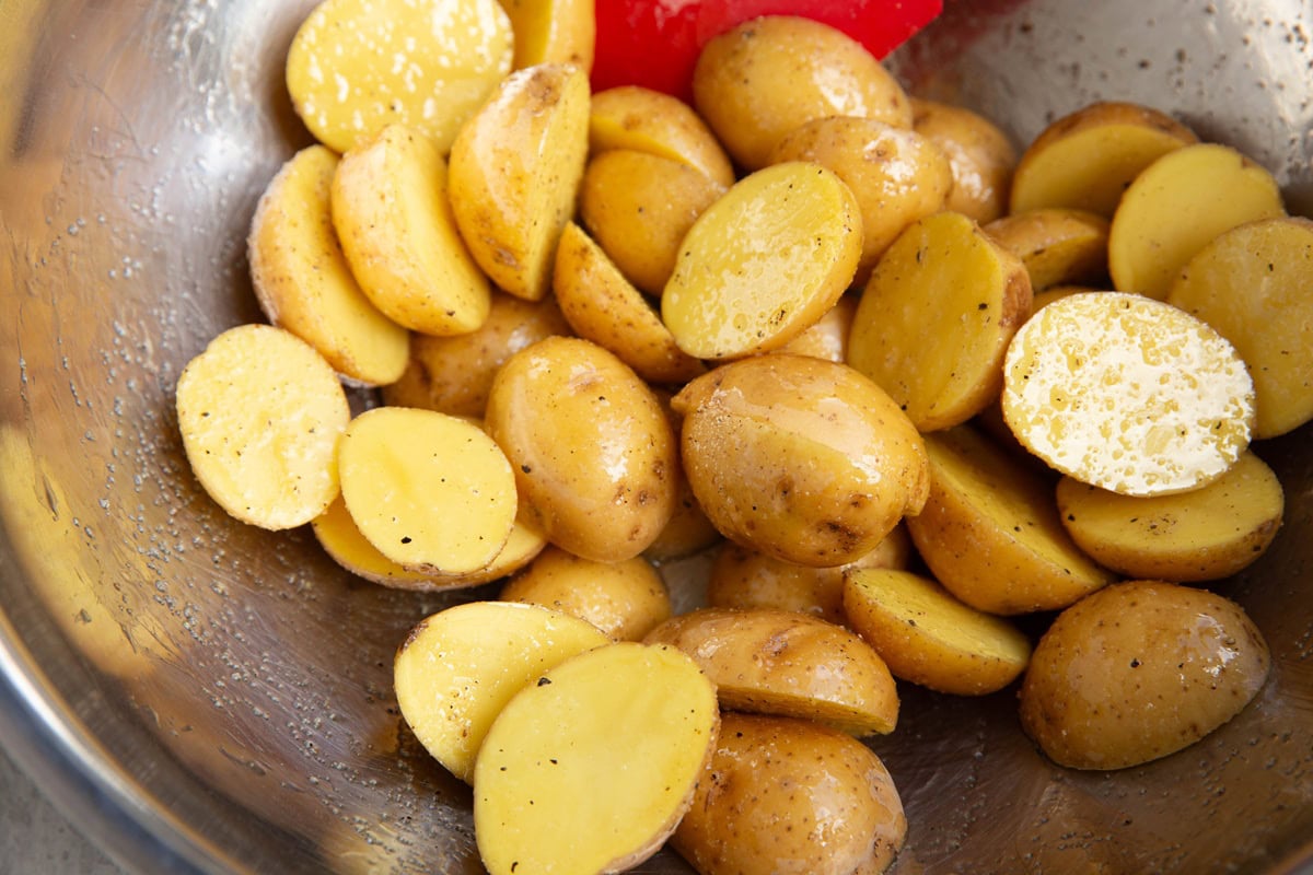 Halved baby potatoes in a large stainless steel bowl that have been tossed in oil and seasonings.
