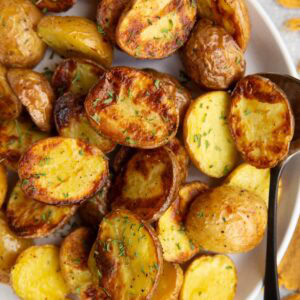 White plate full of golden brown baby potatoes that are sprinkled with dried parsley and a serving spoon is to the side.