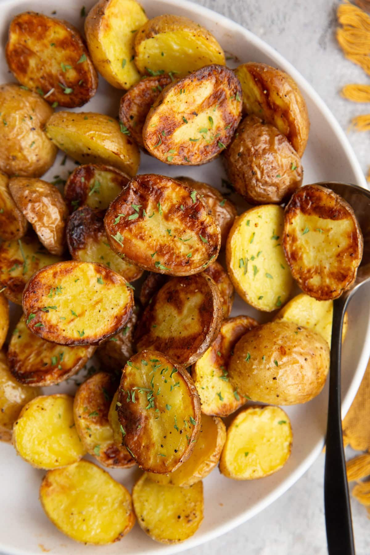 White plate full of golden brown baby potatoes that are sprinkled with dried parsley and a serving spoon is to the side.