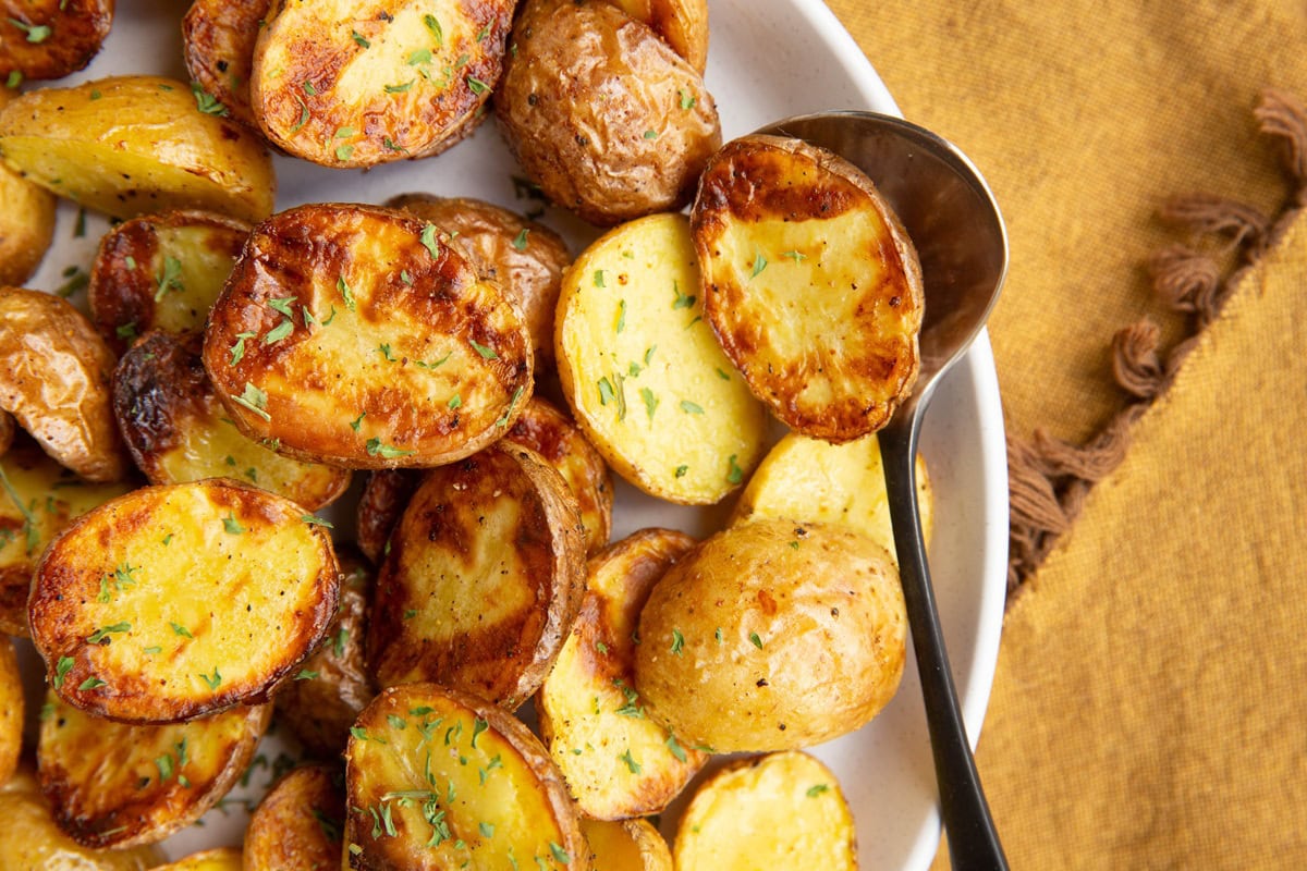 Plate of golden brown baby potatoes with a serving spoon and a golden napkin to the side.