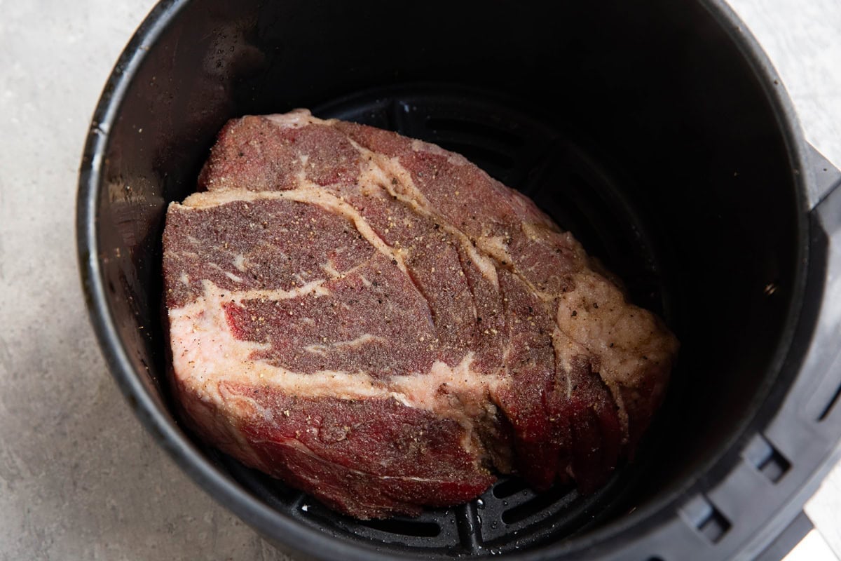 Air fryer basket with a seasoned chuck roast inside, ready to be air fried.