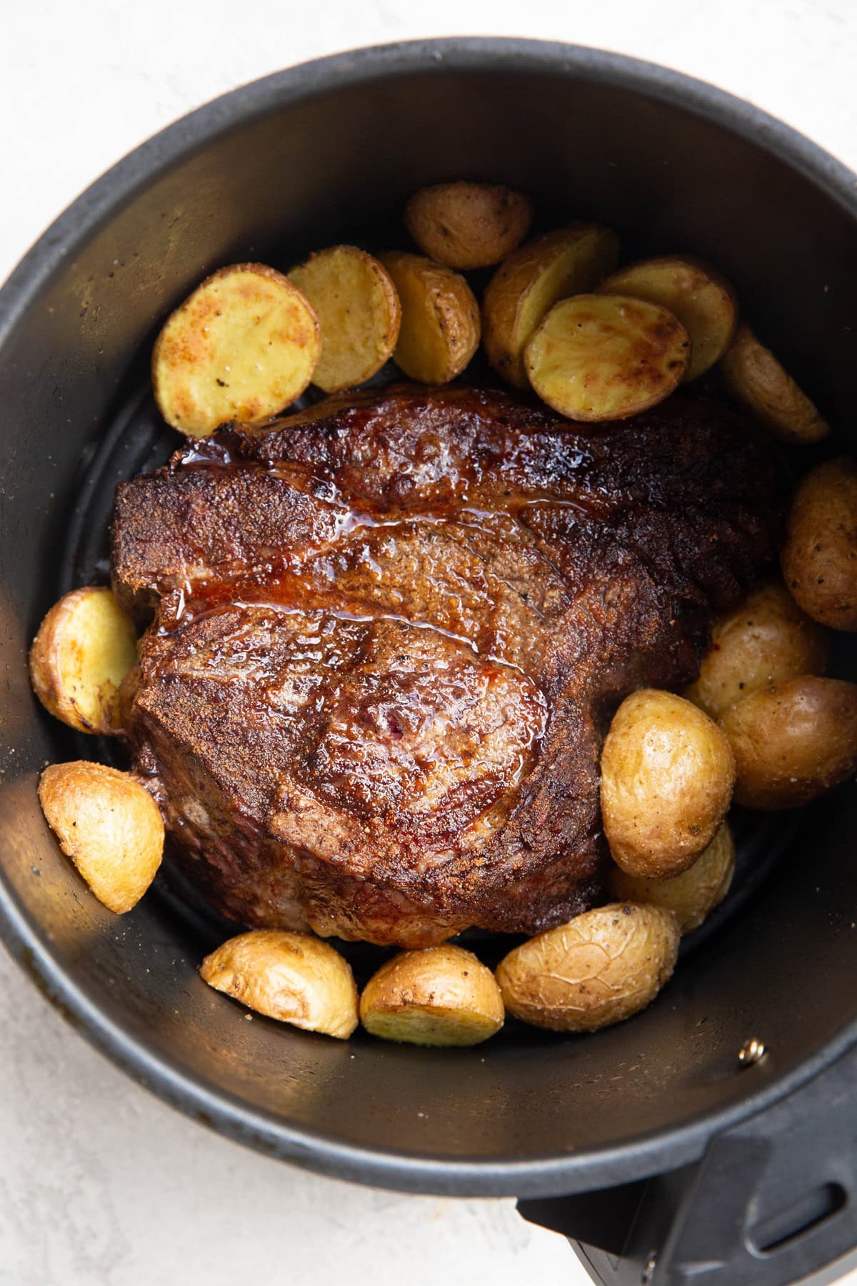 Golden brown cooked chuck roast inside of an air fryer basket with golden brown cooked potatoes surrounding the roast. Ready to serve.