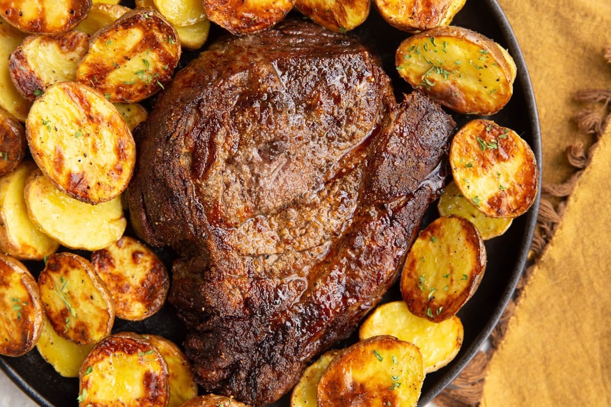 Plate with a whole golden brown cooked chuck roast surrounded in golden-brown cooked baby potatoes. Sprinkled with fresh parsley and a golden napkin to the side.